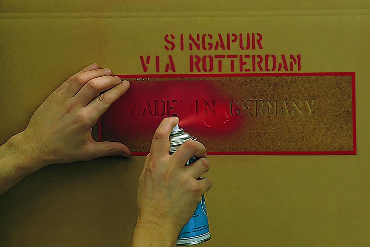 Beschriftung eines Kartons mit Signierspray und Signierschablone Ein Karton wird mit rotem Signierspray aus der Dose und einer Signierschablone beschriftet